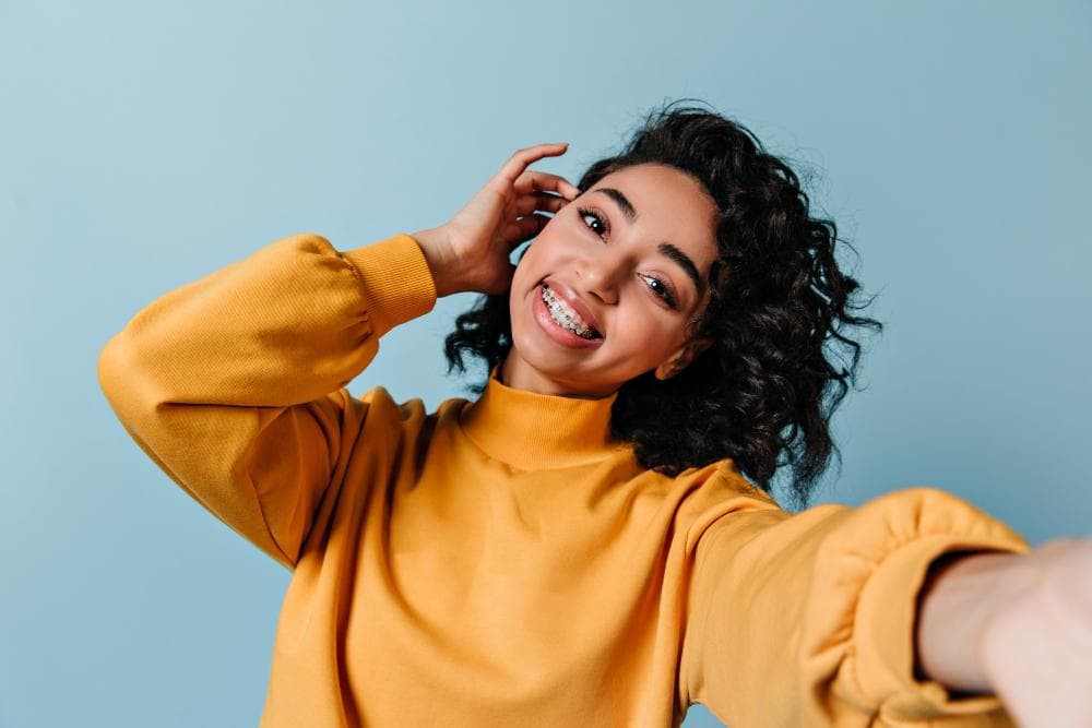 A smiling young woman with curly hair poses for a selfie. She is wearing a bright yellow sweatshirt and has braces on her teeth, showing a confident and cheerful expression. The background is a soft blue.
