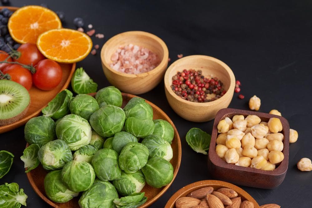 An assortment of fresh vegetables and fruits arranged on a dark surface, featuring Brussels sprouts, tomatoes, blueberries, kiwi, and oranges. There are also small bowls filled with pink Himalayan salt, black peppercorns, and chickpeas.