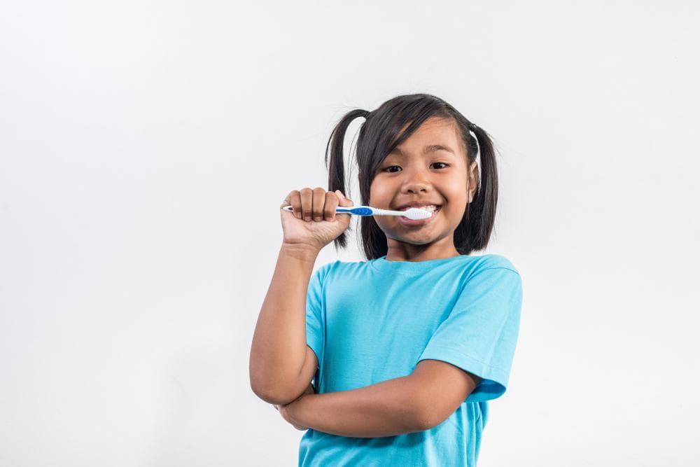 A cheerful young girl with two ponytails is holding a toothbrush with toothpaste on it. She is smiling widely, showing her teeth, and is dressed in a bright blue shirt. The background is plain white.