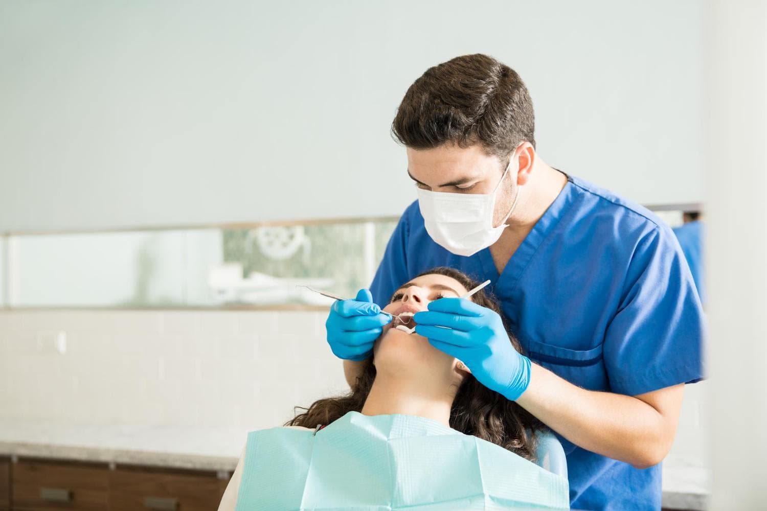 A dentist in blue scrubs and a face mask is examining a female patient's mouth. The patient is reclining back in a dental chair, with her mouth open. The dentist is using dental instruments while focused on the patient's dental health.
