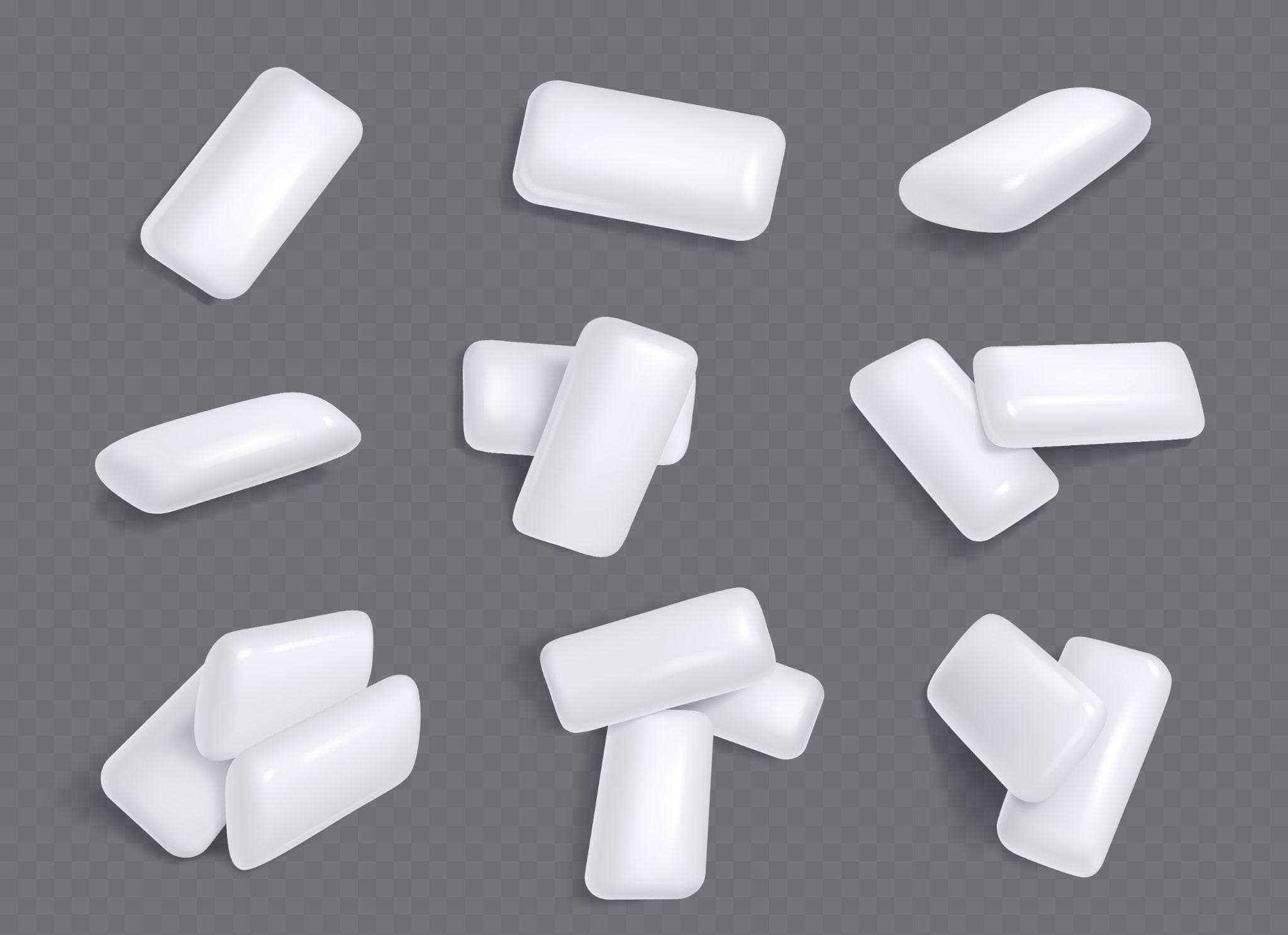 A collection of white chewing gums of different shapes and angles scattered across a gray background. The gums are smooth and shiny, indicating a fresh appearance.
